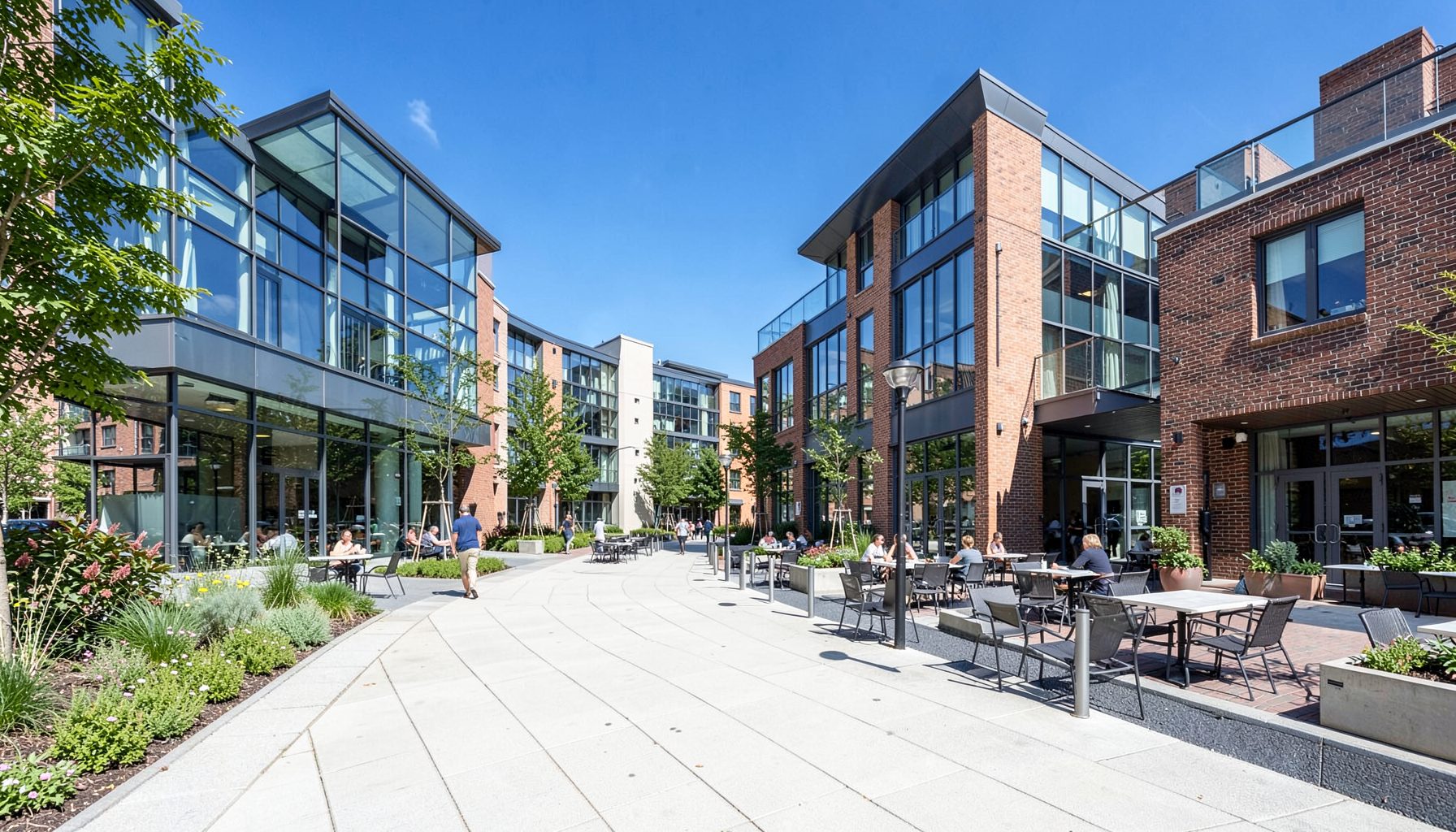 Modern suburban mixed-use development with wide sidewalks, outdoor dining, and contemporary glass-and-brick buildings under clear skies